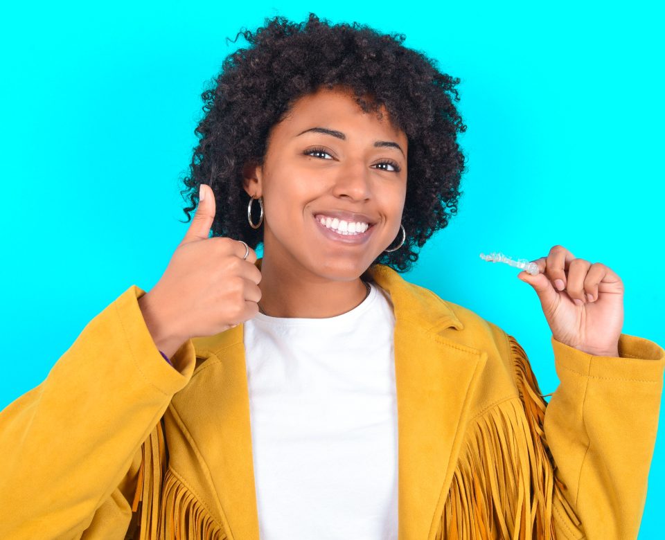 Young Woman With Afro Hairstyle Wearing Yellow Fringe Jacket Over Blue Background Holding An Invisible Braces Aligner And Rising Thumb Up, Recommending This New Treatment. Dental Healthcare Concept.