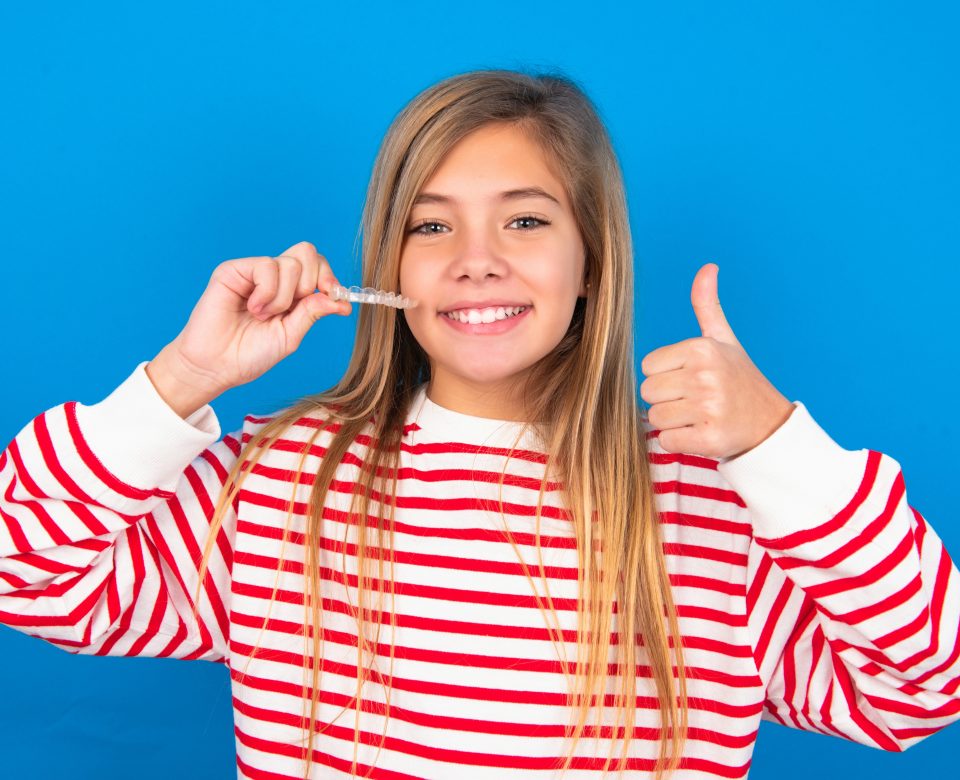 Caucasian Teen Girl Wearing Striped Shirt Over Blue Studio Background Holding An Invisible Braces Aligner And Rising Thumb Up, Recommending This New Treatment. Dental Healthcare Concept.